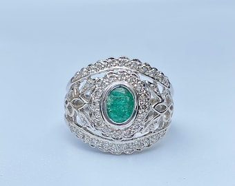 May include: A silver ring featuring a green emerald gemstone surrounded by smaller white gemstones. The ring has a delicate, intricate design.