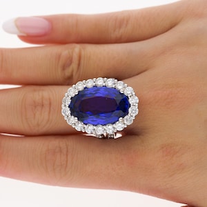 Könnte beinhalten: Ein Ring mit einem großen ovalen blauen Edelstein, umgeben von einem Heiligenschein aus kleineren weißen Edelsteinen.