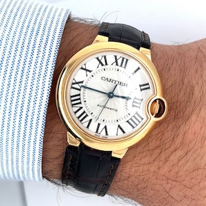 May include: A gold Cartier watch with a round white face and black leather band. The watch face has Roman numerals and the words 'Cartier' and 'Automatic'.