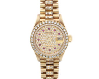 Pode incluir: Um relógio de pulso dourado com moldura e pulseira cravejadas de diamantes. O mostrador do relógio apresenta uma superfície incrustada de diamantes com detalhes de pedras preciosas vermelhas e um mostrador de data. Um item de luxo.
