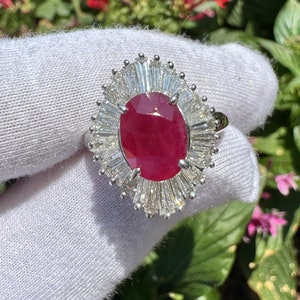 Puede incluir: Un anillo con un gran rubí ovalado rodeado por un halo de diamantes más pequeños. El anillo está engastado en oro blanco.