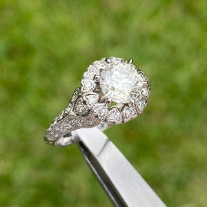 May include: A silver engagement ring with a large, round, clear diamond surrounded by a halo of smaller diamonds. The band is decorated with intricate details.