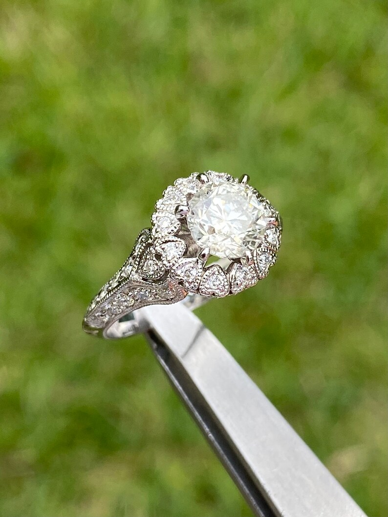 May include: A silver engagement ring with a large, round, clear diamond surrounded by a halo of smaller diamonds. The band is decorated with intricate details.