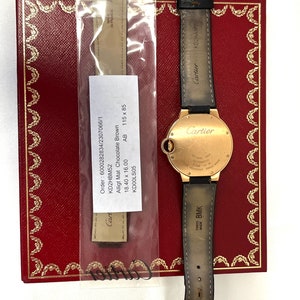 May include: A gold Cartier watch with a brown alligator strap. The watch face is round and has Roman numeral hour markers. The watch is in a box with a red interior. The box has the Cartier logo on it. The watch is also accompanied by a brown leather strap. The strap is labeled 'Cartier' and 'Swiss Made BMK'. The strap is also labeled 'Made in France ANJ'. The watch is labeled 'Cartier' and 'Automatic 300m Water Resistant Swiss Made'. The watch is labeled 'Order: 6000282834/2307066/1 KD2HBM52 Alligt Mat Chocolate Brown 18.40 x 16.00 KD00LS05 AB 115 x 85'.