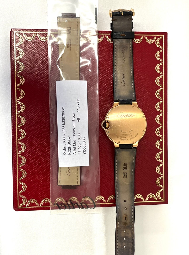 May include: A gold Cartier watch with a brown alligator strap. The watch face is round and has Roman numeral hour markers. The watch is in a box with a red interior. The box has the Cartier logo on it. The watch is also accompanied by a brown leather strap. The strap is labeled 'Cartier' and 'Swiss Made BMK'. The strap is also labeled 'Made in France ANJ'. The watch is labeled 'Cartier' and 'Automatic 300m Water Resistant Swiss Made'. The watch is labeled 'Order: 6000282834/2307066/1 KD2HBM52 Alligt Mat Chocolate Brown 18.40 x 16.00 KD00LS05 AB 115 x 85'.