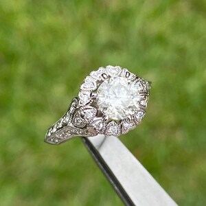 May include: A silver engagement ring with a large, round, clear diamond surrounded by smaller diamonds in a halo setting. The band is decorated with intricate details.