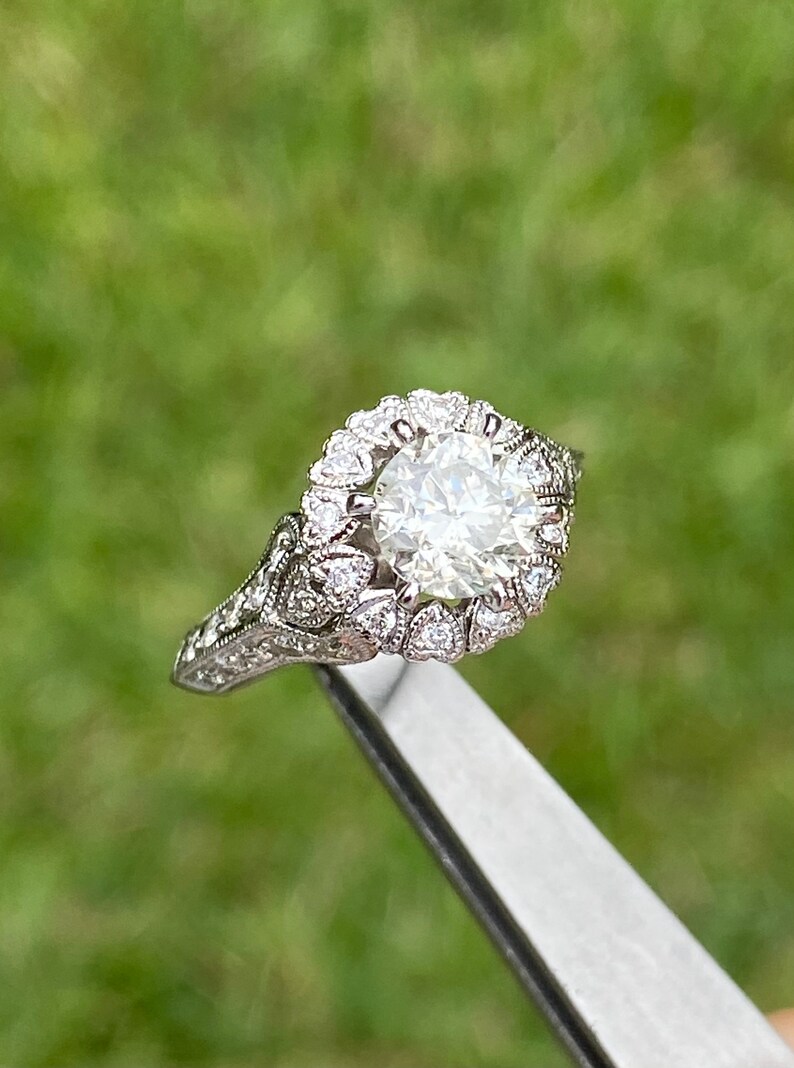 May include: A silver engagement ring with a large, round, clear diamond surrounded by smaller diamonds in a halo setting. The band is decorated with intricate details.