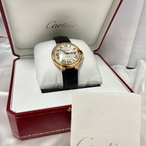 May include: A gold Cartier watch with a white face and black leather band in a red box with the Cartier logo on the lid and a white card with the Cartier logo.