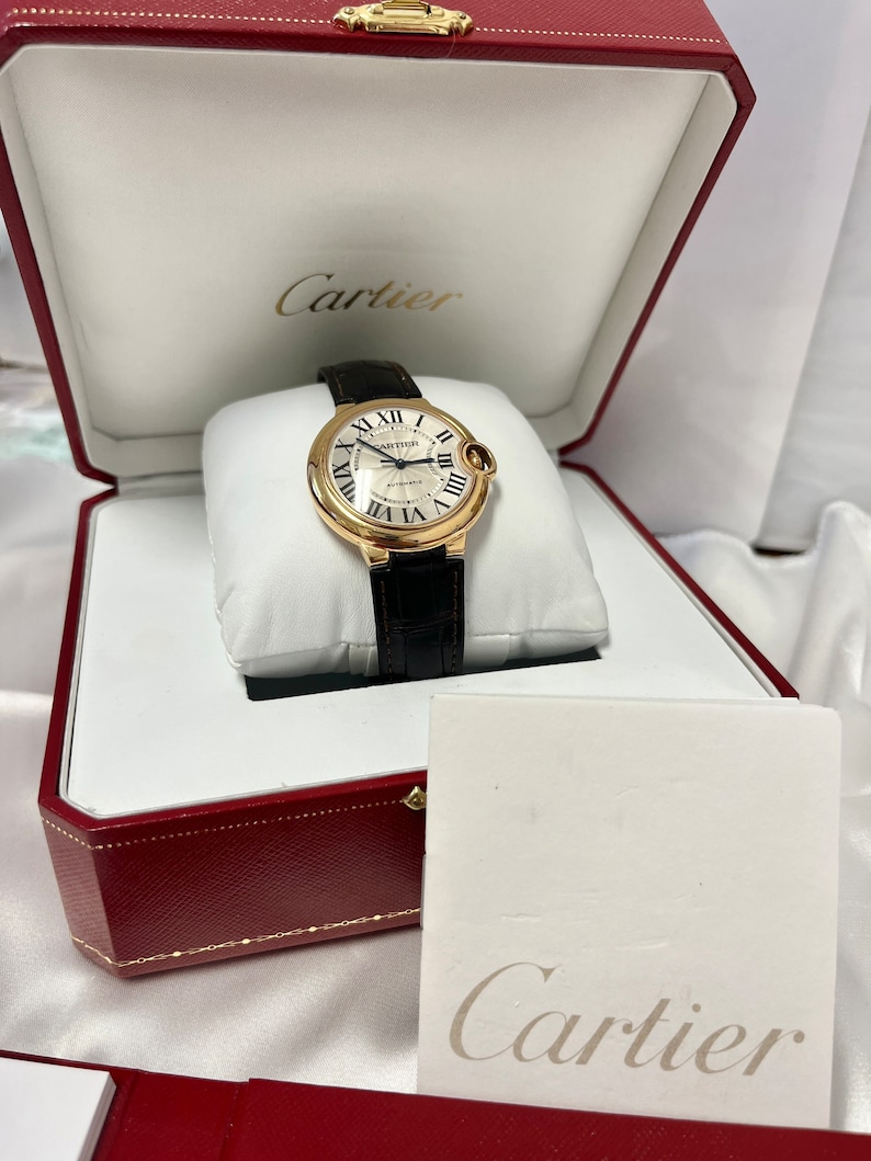 May include: A gold Cartier watch with a white face and black leather band in a red box with the Cartier logo on the lid and a white card with the Cartier logo.