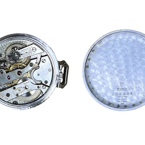 May include: A silver pocket watch with a detailed view of the inner workings of the watch movement. The back of the watch is engraved with the words "Platine" and the number "256991".