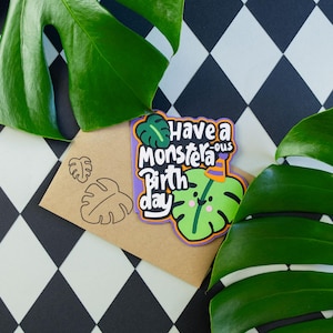 Può includere: Un biglietto di compleanno con un design a foglia di monstera e la scritta "Have a Monster-ous Birthday". Il biglietto è su una busta marrone con timbri a forma di foglia di monstera. Lo sfondo è un motivo a rombi in bianco e nero con foglie di monstera verdi.