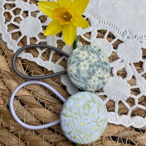 Könnte beinhalten: Zwei Haargummis mit floralen Stoffbezogenen Knöpfen. Die Knöpfe sind hellblau mit einem grünen Blumenmuster und gelben Akzenten. Die Haargummis liegen auf einer gewebten Oberfläche mit einer weißen Spitze.