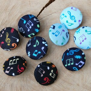 Könnte beinhalten: Neun runde Stoffknöpfe mit schwarzem Hintergrund und bunten Musiknoten darauf gedruckt. Die Knöpfe sind in einem Kreis auf einer Holzoberfläche angeordnet.