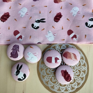 Könnte beinhalten: Sechs runde rosa Stoffknöpfe mit verschiedenen Tierillustrationen. Die Knöpfe zeigen ein Kaninchen, ein Meerschweinchen und einen Igel.