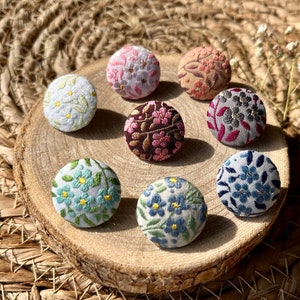 Könnte beinhalten: Eine Sammlung von sieben runden, stoffbezogenen Knöpfen mit verschiedenen Blumenmustern in den Farben Rosa, Blau, Weiß, Grau und Braun. Jeder Knopf zeigt eine aufwendige Stickerei mit metallischen Fadenakzenten, angeordnet in einem Kreis.