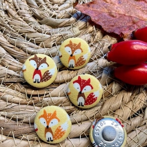 Könnte beinhalten: Vier gelbe Stoffknöpfe mit einem Fuchsdesign. Die Füchse sind orange und weiß mit schwarzen Augen und Nasen. Die Knöpfe befinden sich auf einer gewebten Strohoberfläche mit einem braunen Blatt und roten Beeren im Hintergrund.