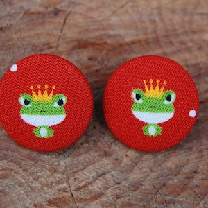 Könnte beinhalten: Ein Paar rote Stoffknopf-Ohrringe mit einem grünen Frosch, der eine goldene Krone trägt.