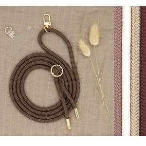 Op de afbeelding: Een bruin gevlochten touw telefoonkoord met gouden hardware en een transparante telefoonhoes. Het koord is opgerold en ligt op een beige stoffen achtergrond.