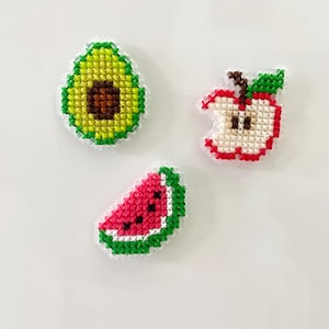 Puede incluir: Tres diseños de frutas en punto de cruz: un aguacate, una manzana mordida y una rodaja de sandía. El aguacate es verde con un hueso marrón. La manzana es roja con un tallo verde. La sandía es rosa con semillas negras y corteza verde.