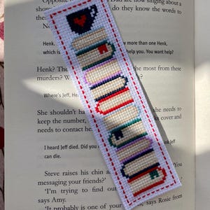 May include: A rectangular white fabric bookmark featuring a cross-stitched design of a stack of books topped with a dark blue mug containing a red heart. The bookmark is bordered by a red stitched outline and rests on an open book page.