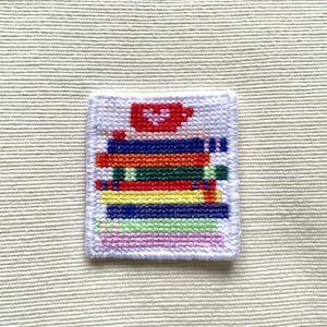 May include: A small, square, embroidered patch featuring a stack of colorful books with a red teacup on top. The books are in shades of blue, orange, green, yellow, and pink, with a white border.