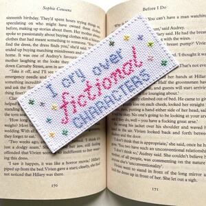 Può includere: Un segnalibro bianco a punto croce con la scritta "I cry over fictional characters" in rosa e blu. Il segnalibro è decorato con piccoli accenti floreali ed è posto su un libro aperto.