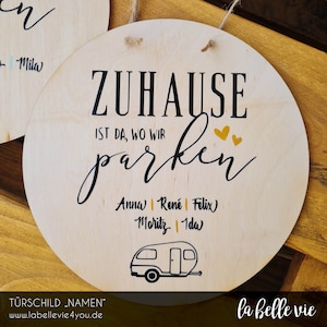 Könnte beinhalten: Ein Holzschild mit dem Text "ZUHAUSE IST DA, WO WIR parken" in schwarzer Schrift. Das Schild zeigt außerdem eine schwarz-weiße Illustration eines Wohnmobils, zwei goldene Herzen und die Namen "Anna", "René", "Felix", "Moritz" und "Ida" in schwarzer Schrift.