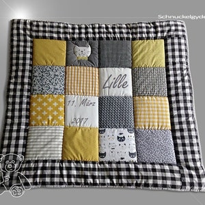 Puede incluir: Una colcha de bebé de patchwork amarillo y gris con un borde a cuadros blanco y negro. La colcha tiene una aplicación de gato, el nombre "Lille" y la fecha "11. März 2017" bordada.