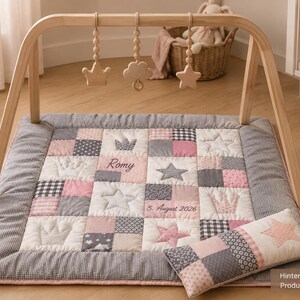 Könnte beinhalten: Ein Baby-Spielbogen mit Holzrahmen und drei hängenden Holzspielzeugen in Kronen-, Wolken- und Sternform. Die quadratische, gesteppte Spielmatte hat ein Patchwork-Design in Rosa, Grau und Weiß, mit gestickten Kronen, Sternen und dem Text Romy 5. August 2026.