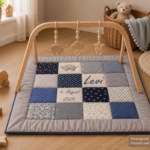Op de afbeelding: Een vierkante patchwork speelmat in blauw-, grijs- en wittinten, voorzien van de naam Levi en de datum 4. August 2026. Het bevat een houten boogframe met drie hangende houten speeltjes in de vorm van een olifant, een wolk en een ster.