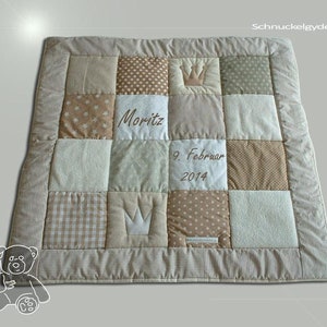 Puede incluir: Una colcha de bebé de patchwork beige y blanco con una corona, el nombre Moritz y la fecha 9 de febrero de 2014 bordada en ella. La colcha está hecha de diferentes telas, incluyendo vichy, lunares y colores sólidos.