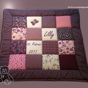 May include: A purple and pink patchwork baby quilt with a butterfly applique and the name "Lilly" embroidered on it. The quilt also has the date "20. Februar 2017" embroidered on it.