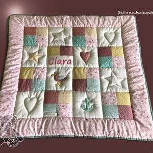 Könnte beinhalten: Eine rosa-grüne Patchwork-Babydecke mit weißem Rand. Die Decke hat verschiedene Applikationen, darunter Sterne, Herzen, einen Vogel und einen Anker. Der Name "Clara" ist in rosa auf die Decke gestickt.