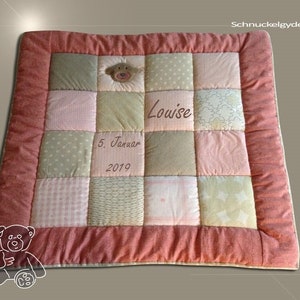 Op de afbeelding: Een patchwork babydeken met een teddybeer applicatie en een roze rand. De deken heeft vierkanten van verschillende pastelstoffen, met de naam "Louise" en de datum "5. Januar 2019" erop geborduurd. De deken is rechthoekig.