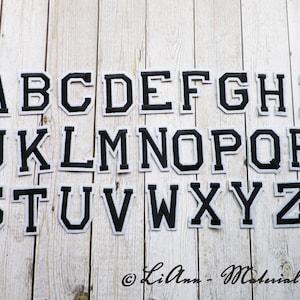 Könnte beinhalten: Schwarze Filzbuchstaben in einer Schriftart im College-Stil. Die Buchstaben sind in alphabetischer Reihenfolge auf einem weißen Holzgrund angeordnet.