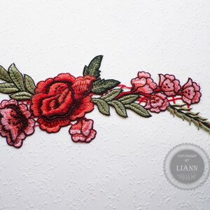 Könnte beinhalten: Gestickte Blumenapplikation mit einer großen roten Rose und kleineren rosa Rosen, grünen Blättern und einem rot-weiß karierten Muster. Die Applikation befindet sich auf einem weißen Hintergrund. Copyright by Liann.