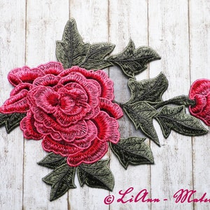 Könnte beinhalten: Eine rosa und grüne gestickte Blumenapplikation mit einer einzigen großen Blume und einer kleineren Blume an einem Stiel. Die Applikation befindet sich auf einem weißen Holzgrund.