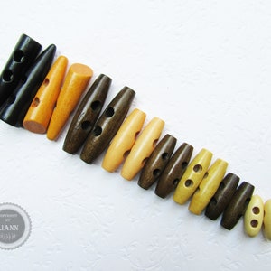 Könnte beinhalten: Eine Sammlung von Holz-Toggle-Knöpfen in verschiedenen Brauntönen, Gelb- und Orangetönen. Die Knöpfe haben zwei Löcher und sind in einer Reihe vom größten zum kleinsten angeordnet.