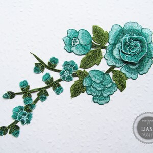 Könnte beinhalten: Tealblaue gestickte Blumenapplikation mit grünen Blättern. Die Applikation zeigt eine große Rose und kleinere Blumen.
