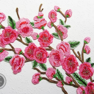 Könnte beinhalten: Rosa bestickte Blumenapplikation mit braunen Stielen und grünen Blättern. Die Applikation zeigt eine Ansammlung von rosa Blüten mit einem zarten Design.