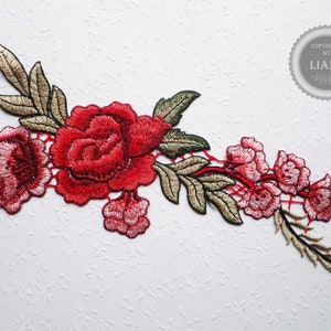 Könnte beinhalten: Rote gestickte Blumenapplikation mit grünen Blättern und einem zarten Spitzenmuster. Die Applikation zeigt eine große Rose und kleinere Rosen mit einem rankenartigen Stiel.