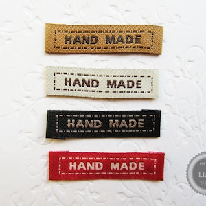 Könnte beinhalten: Vier Stoff-Etiketten mit dem Text "HAND MADE" in brauner Schrift. Die Etiketten sind in Braun, Weiß, Schwarz und Rot.