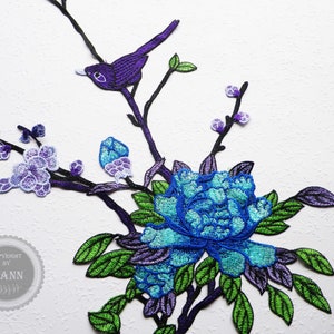 Könnte beinhalten: Violette und blaue gestickte Applikation mit einem Vogel, der auf einem Zweig mit Blumen und Blättern sitzt. Die Applikation ist perfekt, um Kleidung, Taschen oder Heimtextilien einen Hauch von Eleganz zu verleihen.