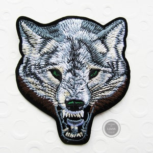 Könnte beinhalten: Gestickter Aufnäher mit dem Kopf eines knurrenden Wolfes mit grünen Augen. Der Wolf ist weiß mit braunem Fell um die Schnauze. Der Aufnäher ist schwarz und hat einen weißen Rand.