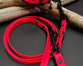 May include: A neon pink dog collar and matching leash set featuring matte black hardware. The collar has a buckle closure and a D-ring, while the leash includes a swivel snap hook and additional rings. Both items are made of a smooth, flexible material.
