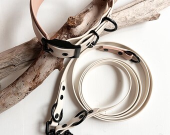 May include: A light beige dog collar and a matching long leash featuring matte black metal hardware, including sturdy D-rings and swivel snap hooks. The collar has a smooth, wide band design with a black buckle, while the leash is coiled.