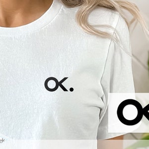 Könnte beinhalten: Weißes T-Shirt mit einem schwarzen Logo, das einen Kreis, ein stilisiertes 'K' und einen Punkt zeigt. Das Logo befindet sich mittig auf der Brust. Ein kleineres Logo ist in der unteren rechten Ecke zu sehen.