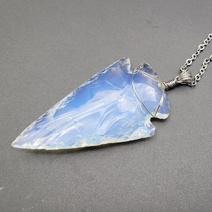 Peut inclure: Un collier avec un pendentif en forme de pointe de flèche opalescente. Le pendentif, de couleur bleu clair, est enveloppé de fil de fer et suspendu à une chaîne argentée. L'ensemble est posé sur un fond sombre.