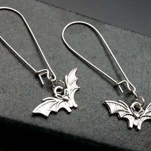 May include: A pair of silver dangle earrings featuring a bat charm. The earrings have a simple wire loop design.