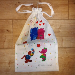 Puede incluir: Bolsa blanca de cordón con una ilustración colorida de elefantes y confeti. La bolsa tiene el texto "© Janine Ullrich" en la parte inferior.